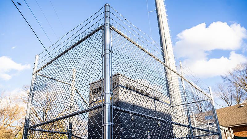Chain-link fence enclosing dark electrical equipment.  A utility pole rises nearby against a blue sky with scattered clouds.