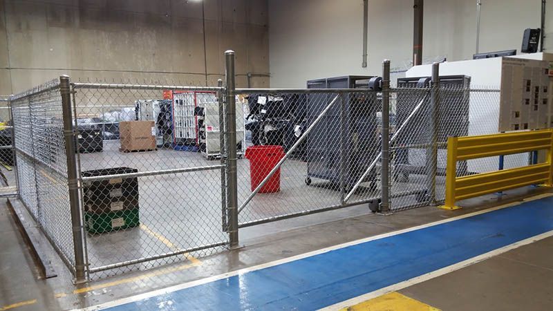 A chain-link fenced area in a warehouse, containing bins, carts, and a trash can. A blue and yellow floor marking is visible.