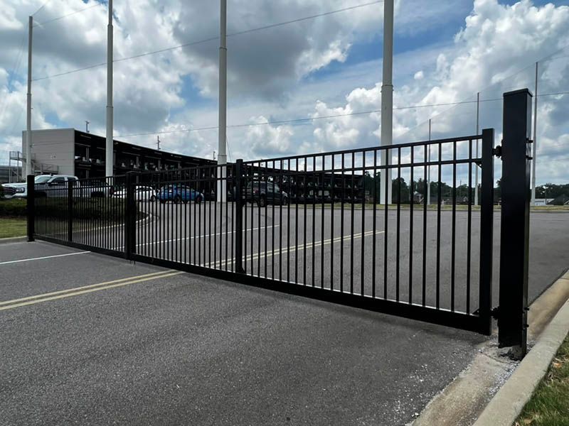 Black metal security gate across a paved area, with a building and golf course poles in the background.