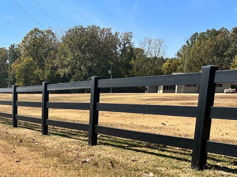 Black wooden fence in a field, trees and a building in the background under a blue sky.