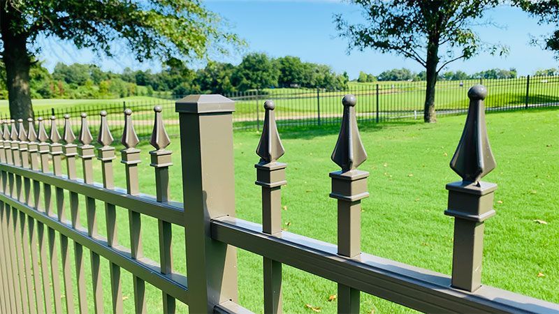A decorative brown metal fence with pointed finials borders a grassy area, with trees and a blue sky in the background.