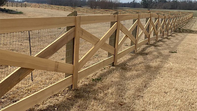 Wooden fence with crisscross design, running along a field of dry grass under a sunny sky.
