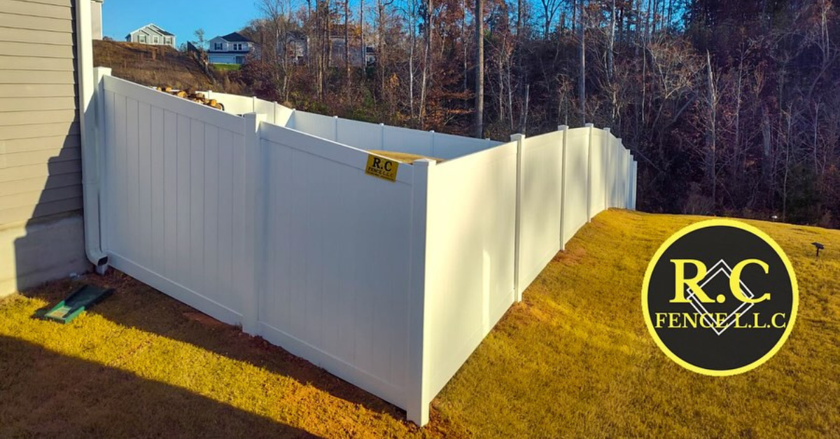 White vinyl fence enclosing a yard on a sloped lawn. Trees are in the background. Company logo in the bottom right corner.