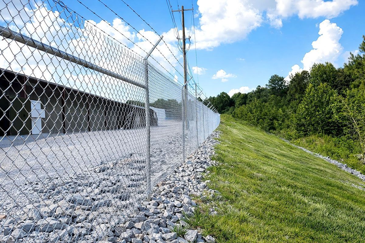 A chain-link fence topped with barbed wire runs beside a gravel area and a grassy embankment under a blue, cloudy sky.