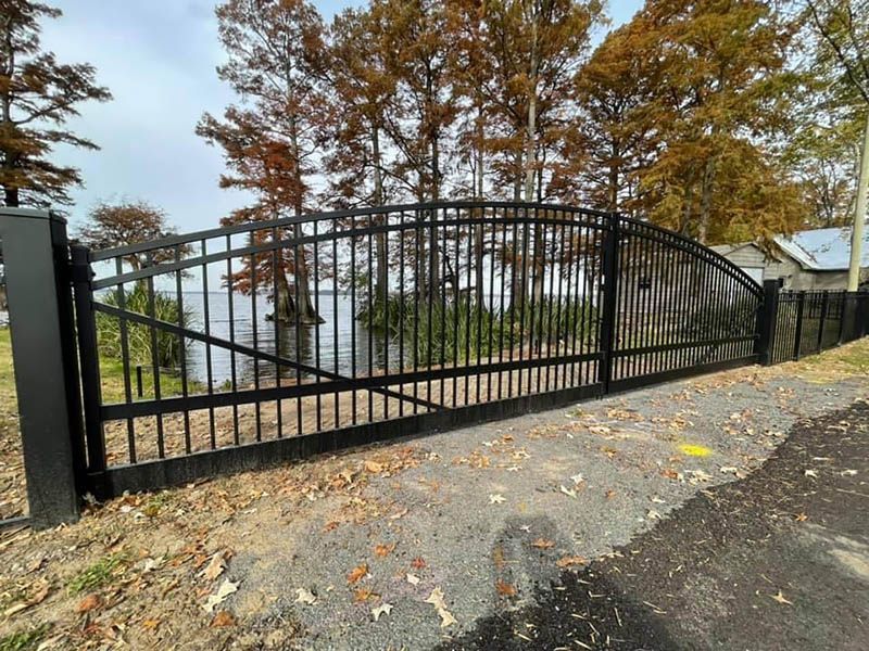 Black metal gate on a gravel driveway, overlooking a body of water with trees in the background.