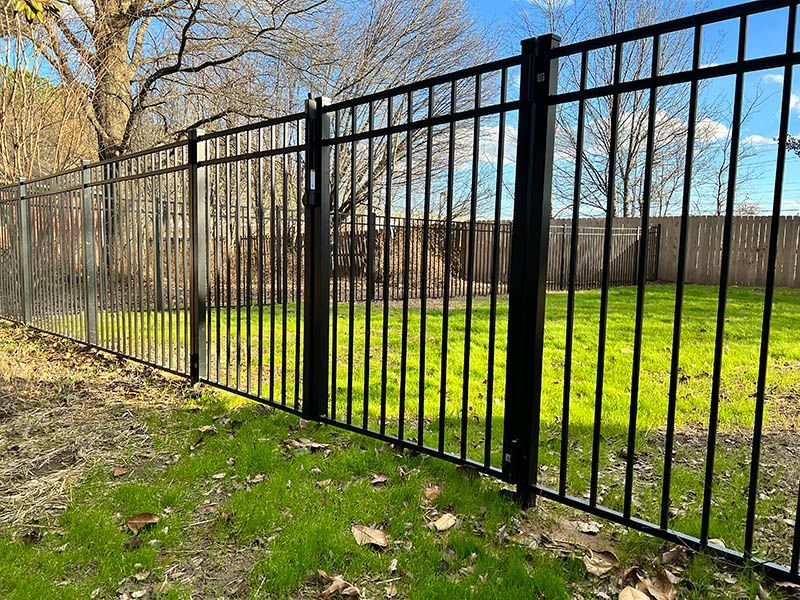 Black metal fence on green grass under a blue sky. The fence separates a yard from a wooden fence in the background.