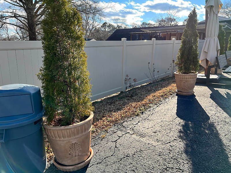 Two potted evergreen trees flank a white fence. The sun casts a shadow across the asphalt driveway. A blue trash can is in the foreground.