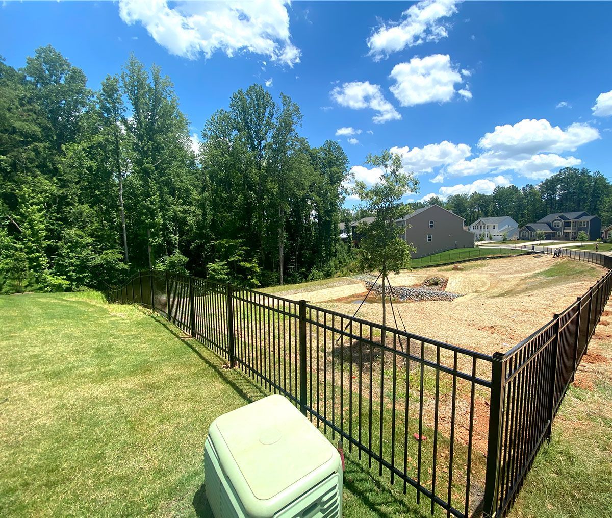 Backyard view with a black fence surrounding a dirt area, with trees in the background, and a blue sky with clouds.