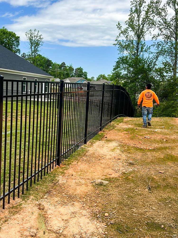 A man walks along a black metal fence, likely inspecting it. The fence borders a grassy yard and a dirt path.