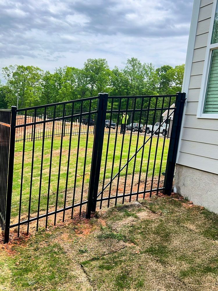 Black metal fence against a tan house. The fence encloses a grassy area with trees in the background and a cloudy sky.