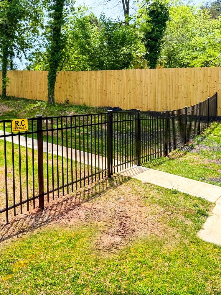 Black metal fence with a light brown wooden fence in the background, separating a grassy yard from a wooded area.