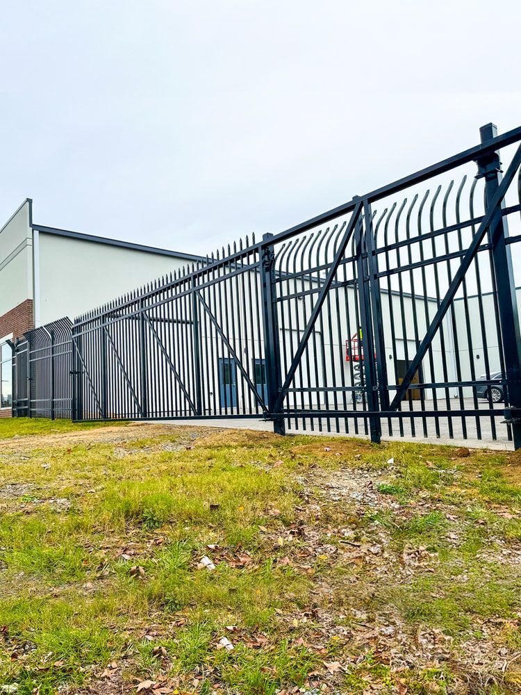Black metal security fence surrounding a commercial building, set on a grassy patch under an overcast sky.