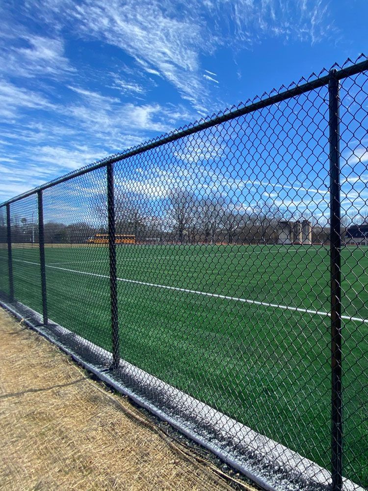 A black chain-link fence surrounds a green turf field under a blue sky with wispy clouds.
