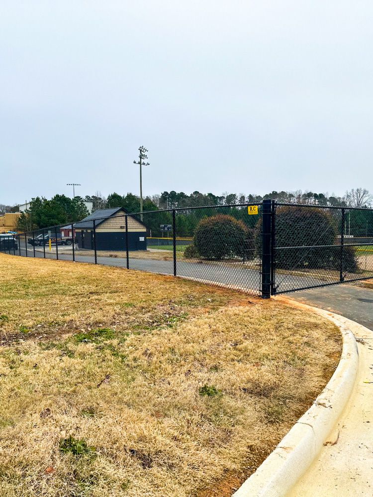 A chain-link fence with a gate surrounds a park area. A small building and trees are visible in the background under an overcast sky.