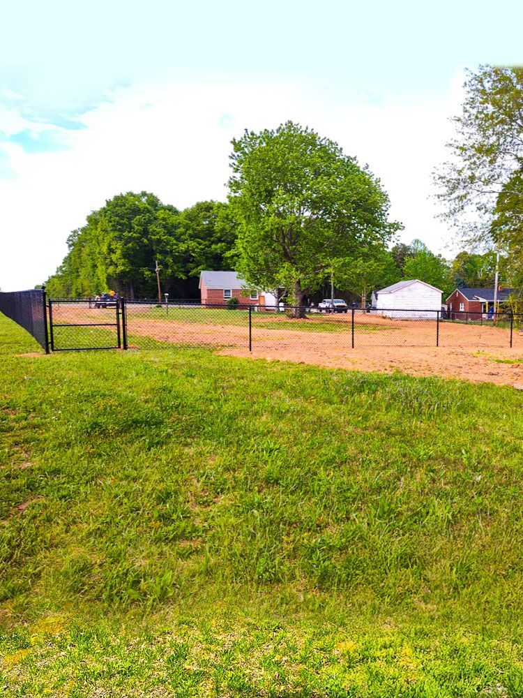 Grassy foreground leads to a dirt path, fencing, trees, and a few houses under a cloudy sky.