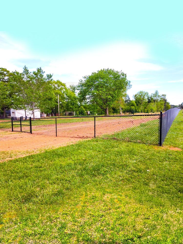 A fenced-in dog park with a dirt area and green grass. Trees and a blue sky are in the background.