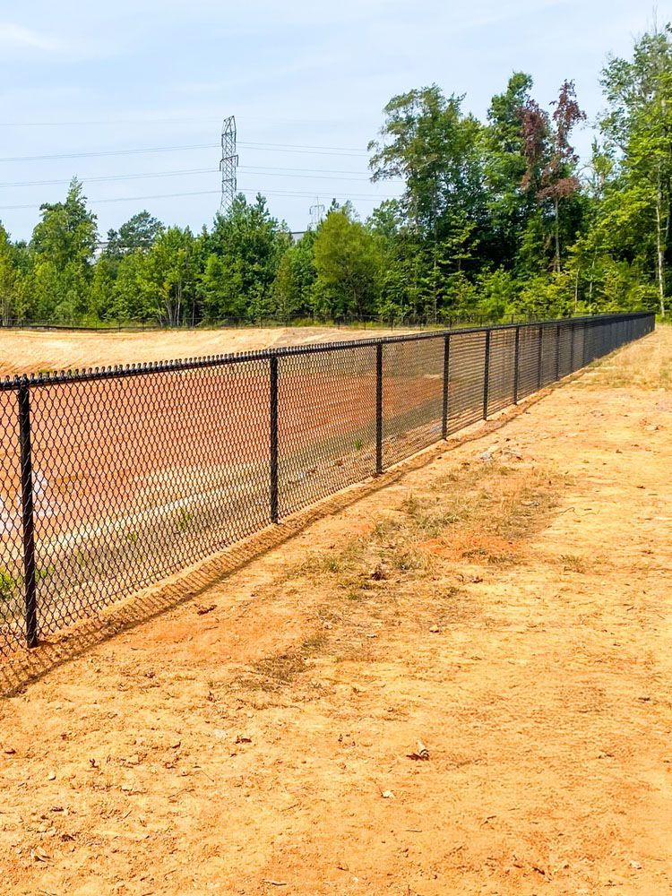 Black chain-link fence bordering a dirt field, trees in the background, and a utility tower in the distance.