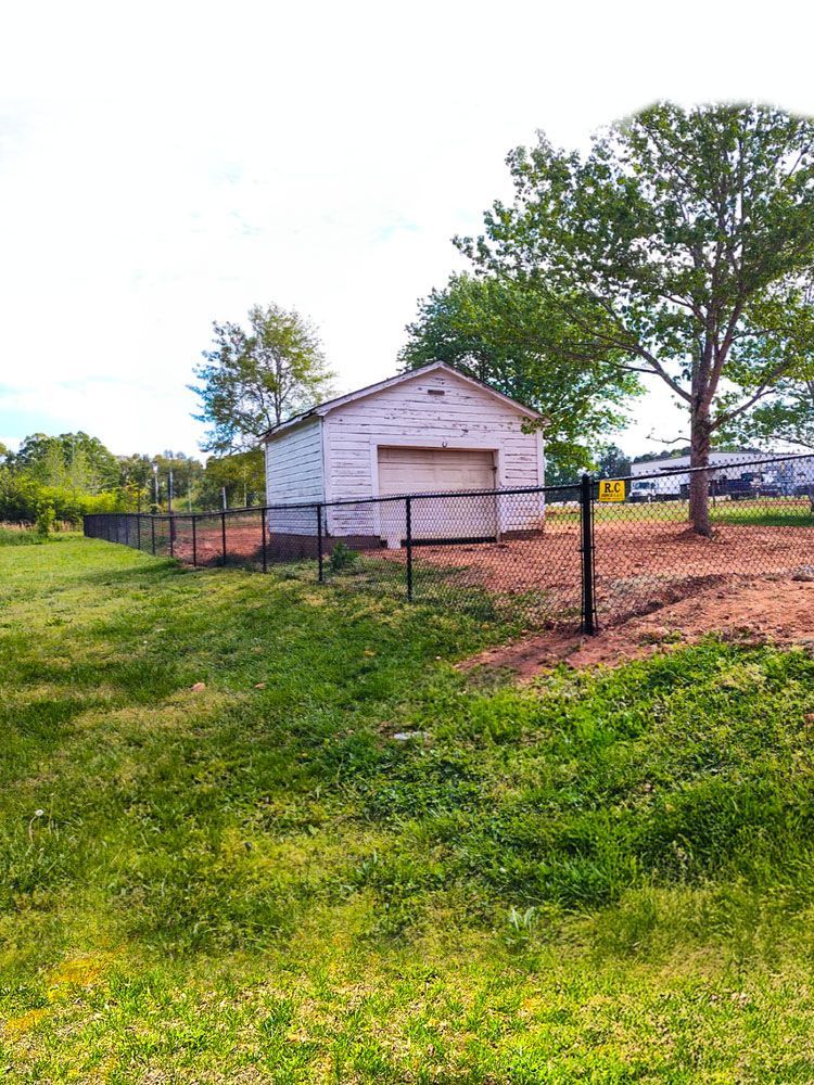 A weathered, white shed with a closed garage door sits behind a black fence on a grassy patch of land.