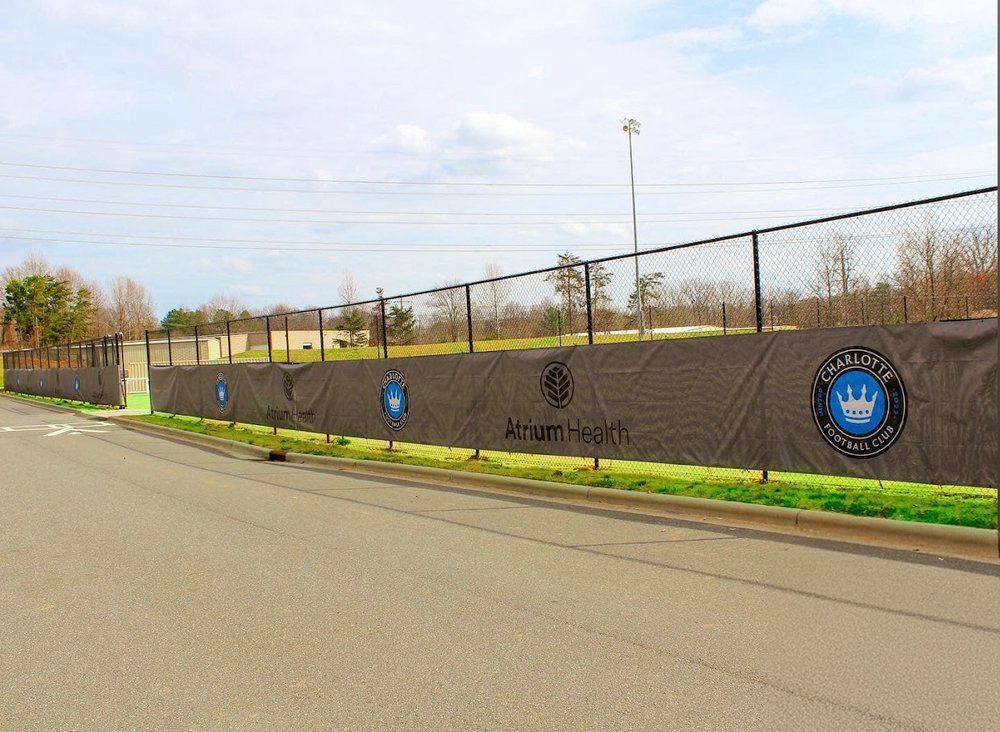Black chain-link fence with gray banner displaying Charlotte FC logos along a road with green grass.