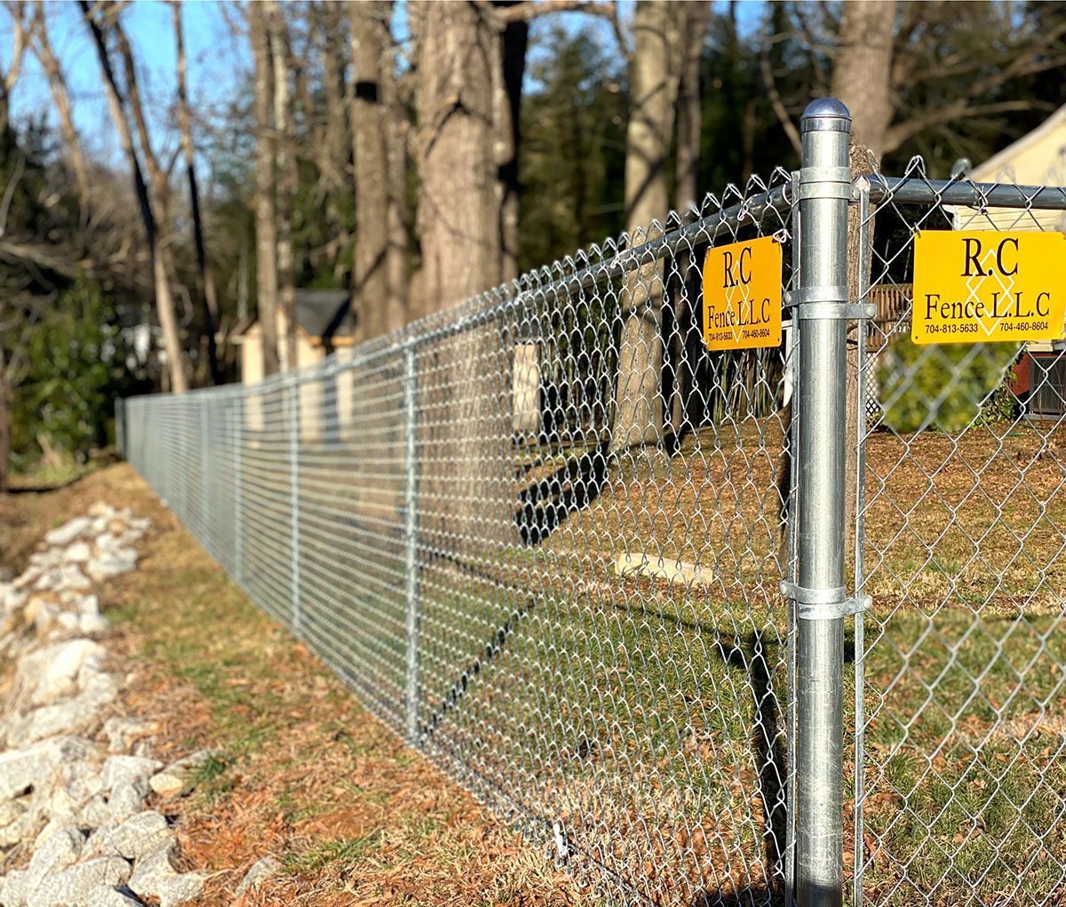 Chain-link fence with a row of rocks on the side, labeled with RC Face LLC, in front of a tree-filled background.