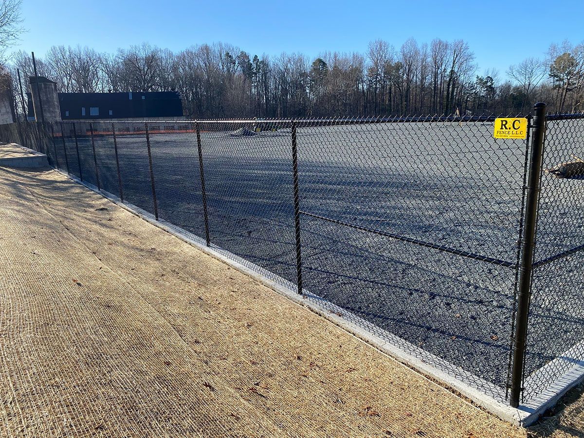 Black chain-link fence with dark netting and a concrete base, set in a field. A small building and trees are in the background.