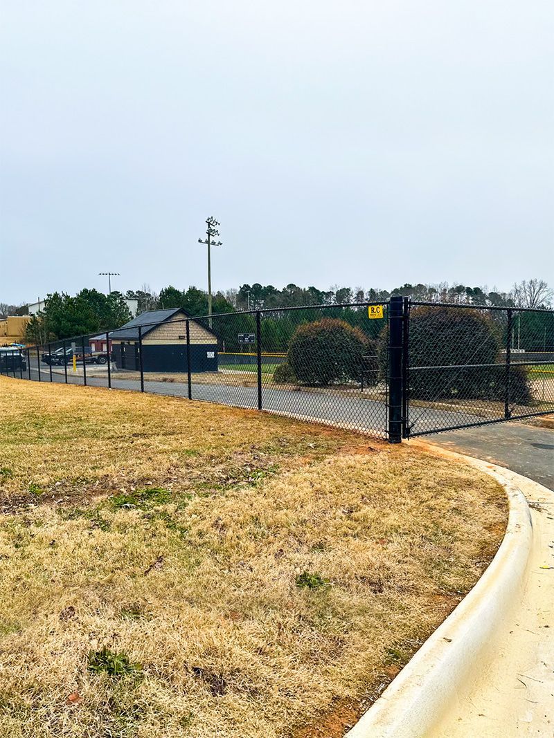 A chain-link fence encloses a grassy area with a small building and trees. A paved road curves in the foreground.
