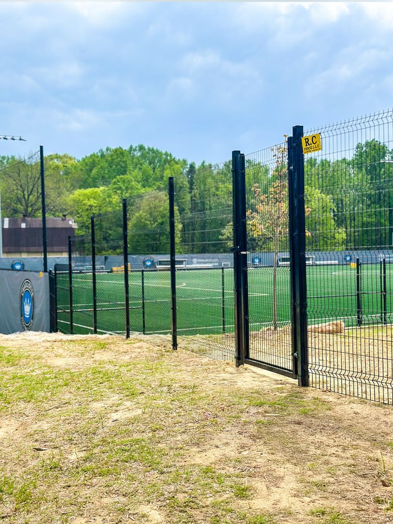 A fenced-in outdoor dog park with black metal posts and artificial turf. A gate is open; trees are in the background.
