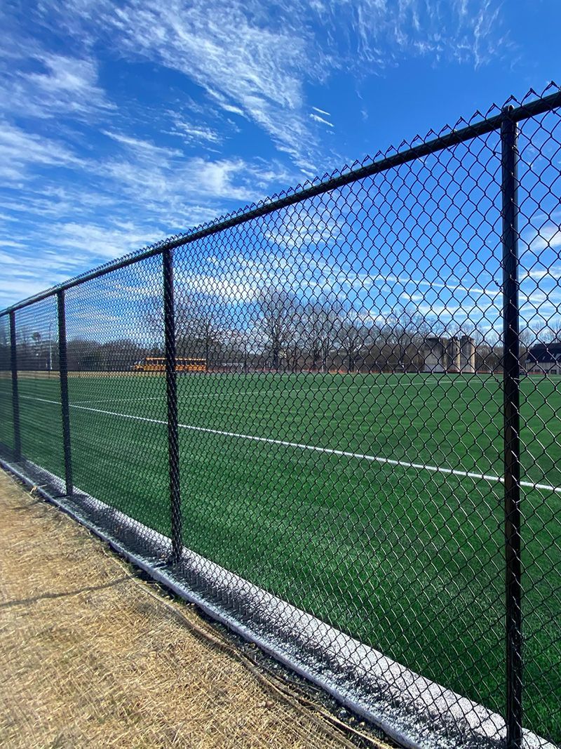 Black chain-link fence bordering a green athletic field under a blue sky with wispy clouds.