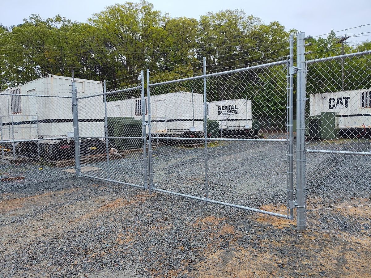 A chain-link fence encloses a gravel yard with white storage containers. Green trees are in the background.