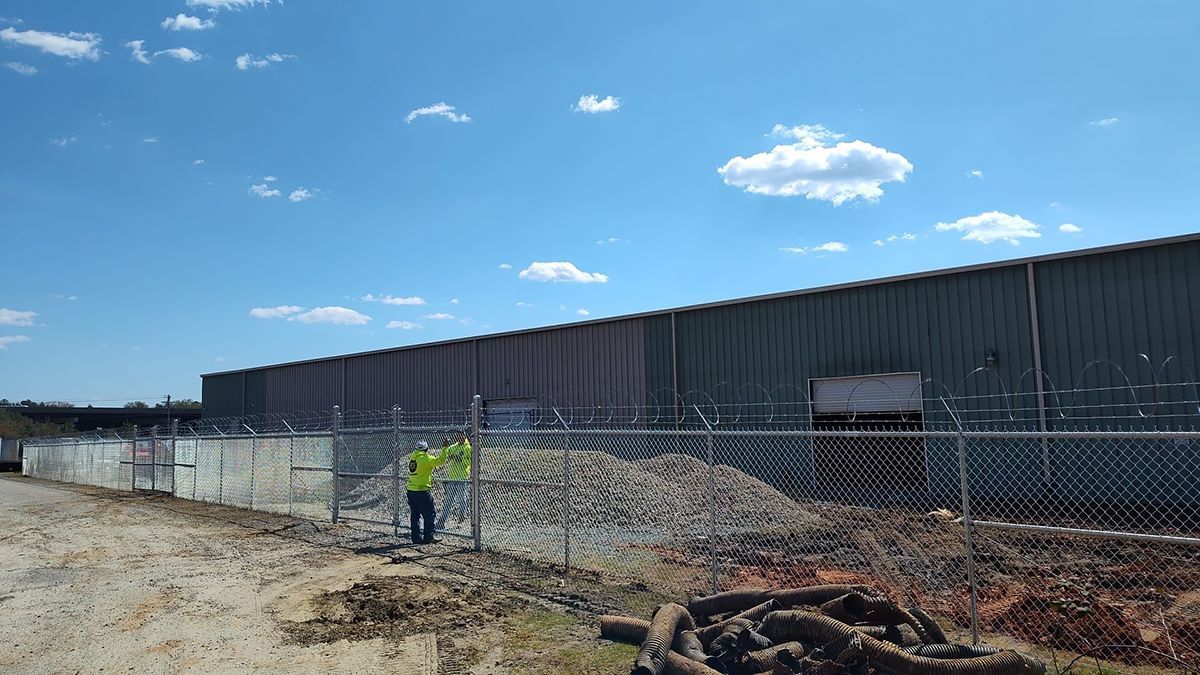 A worker in a neon vest near a chain-link fence in front of a long industrial building on a sunny day.