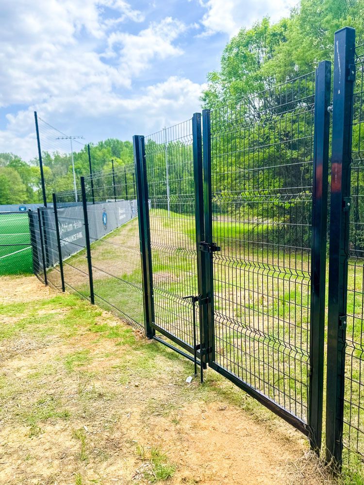 A black metal gate open in a fence, with a soccer field in the background. Green grass and trees surround the field.