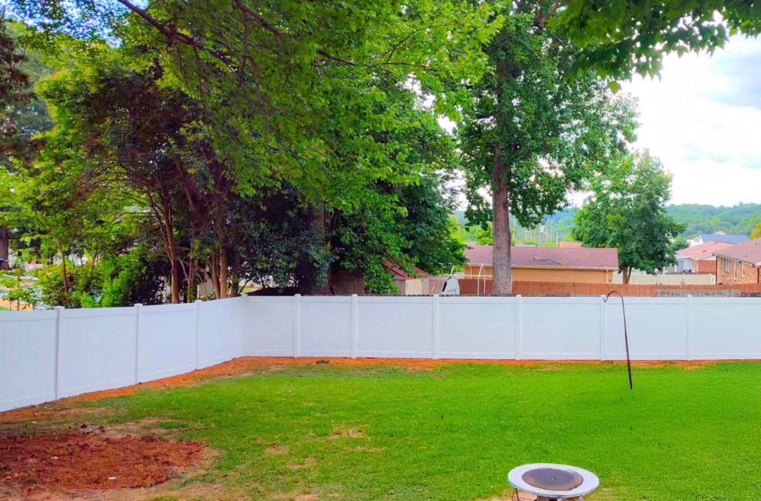 Lush green backyard bordered by a white fence, with trees in the background, and a small fire pit in the foreground.