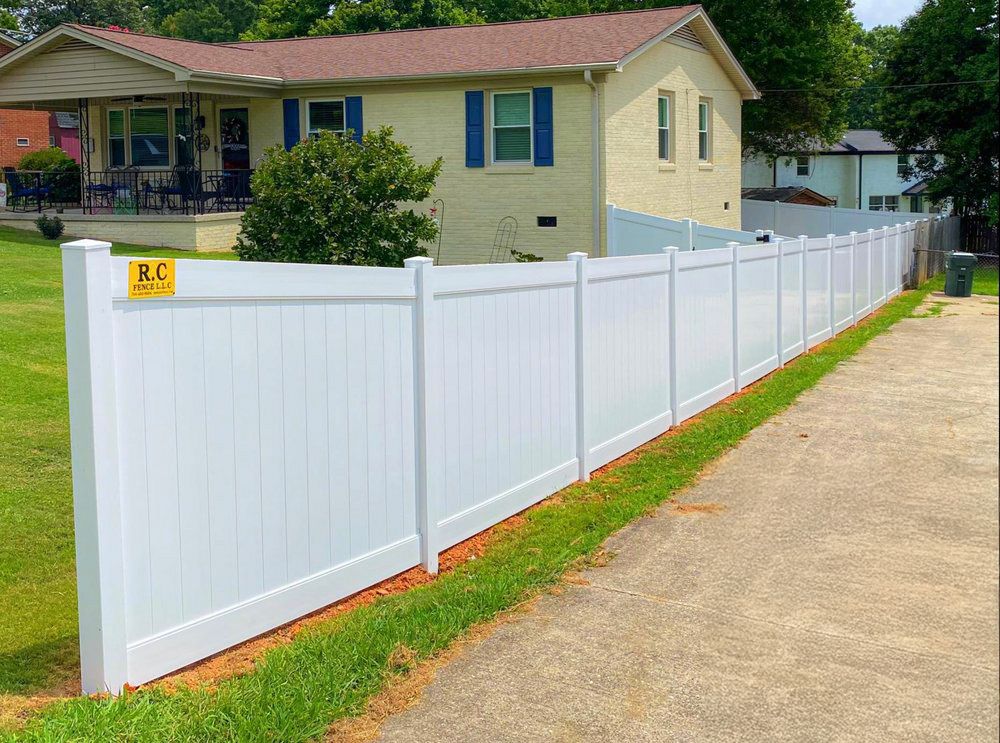 White vinyl fence along a driveway, next to a two-story yellow house with blue shutters. Green grass borders the fence.