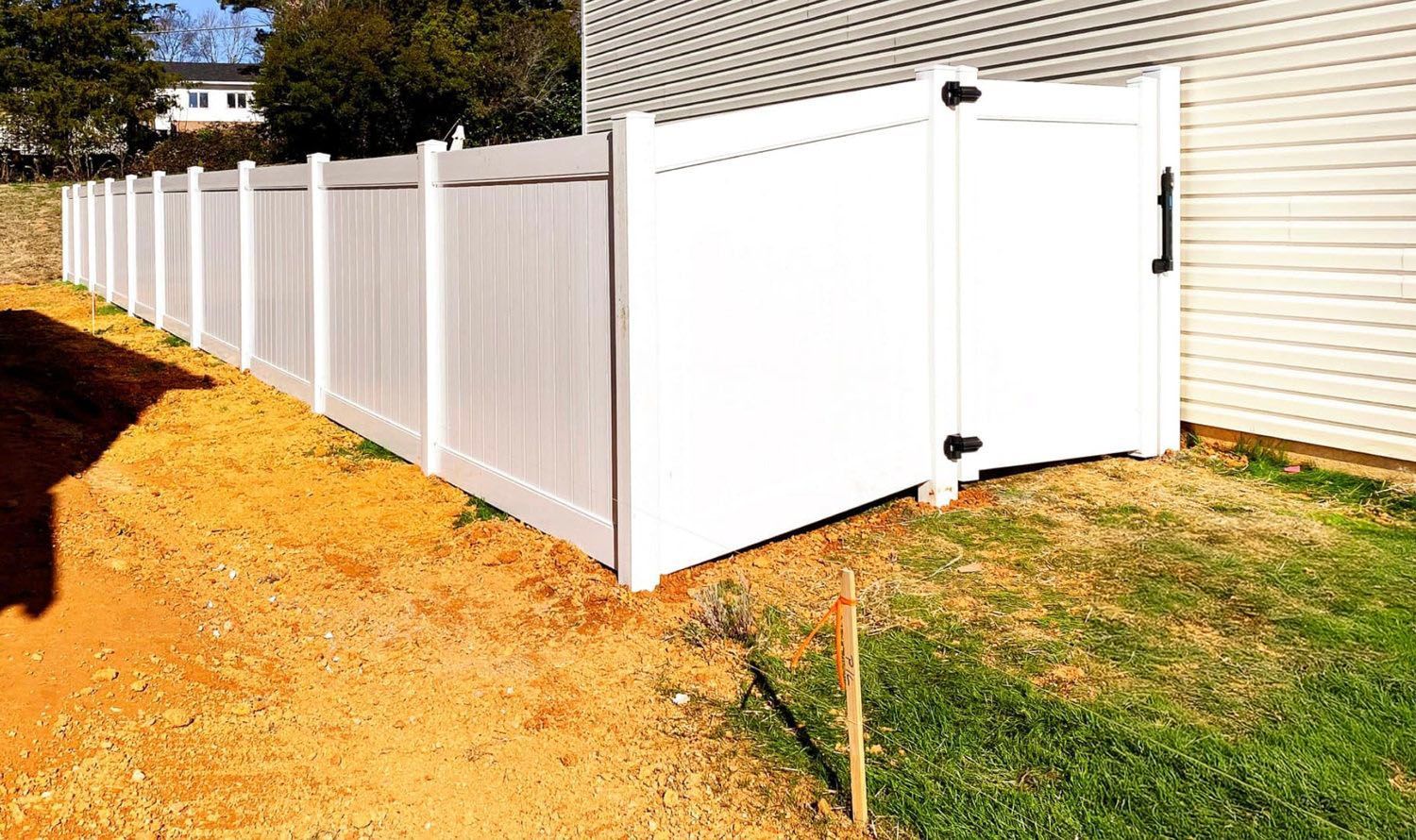 White vinyl fence with a gate next to a building, on a sunny day. Dirt and grass are present.