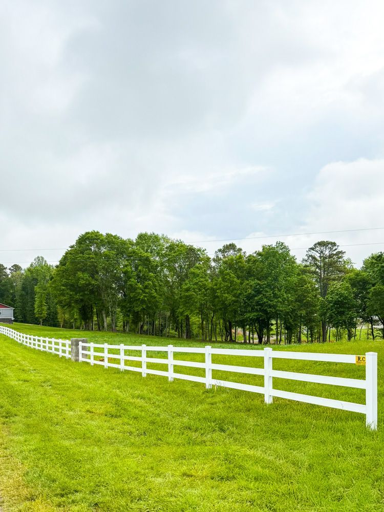 White fence bordering a grassy field with trees in the background under a cloudy sky.