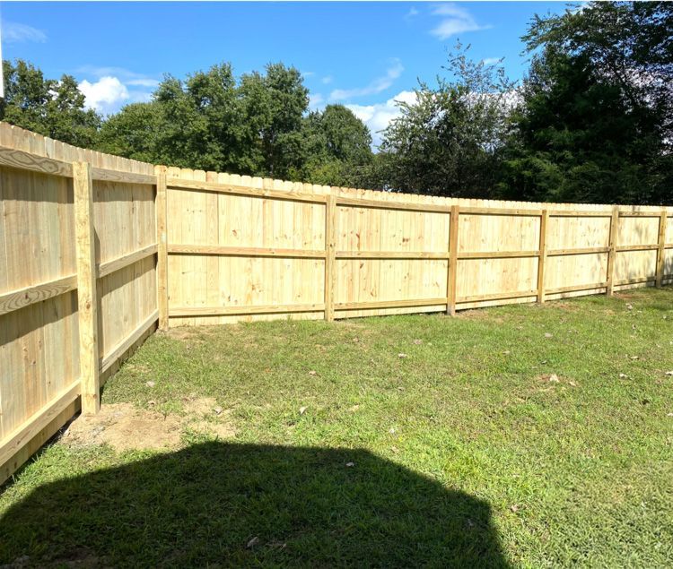 Wooden fence enclosing a grassy backyard on a sunny day. The fence is light-colored and borders the yard, with trees visible in the background.