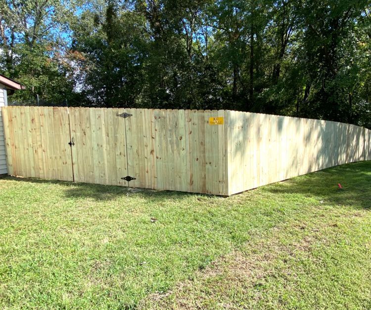 A new wooden fence encloses a grassy yard. A gate is on the left. Trees and a house are in the background.