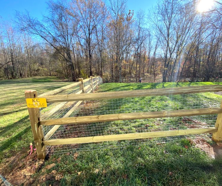 A wooden split-rail fence with wire mesh spans a grassy area, with trees in the background under a blue sky.