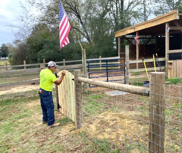 A man in a neon shirt installs a wooden gate on a farm. An American flag flies in the background, and a small shed is visible.