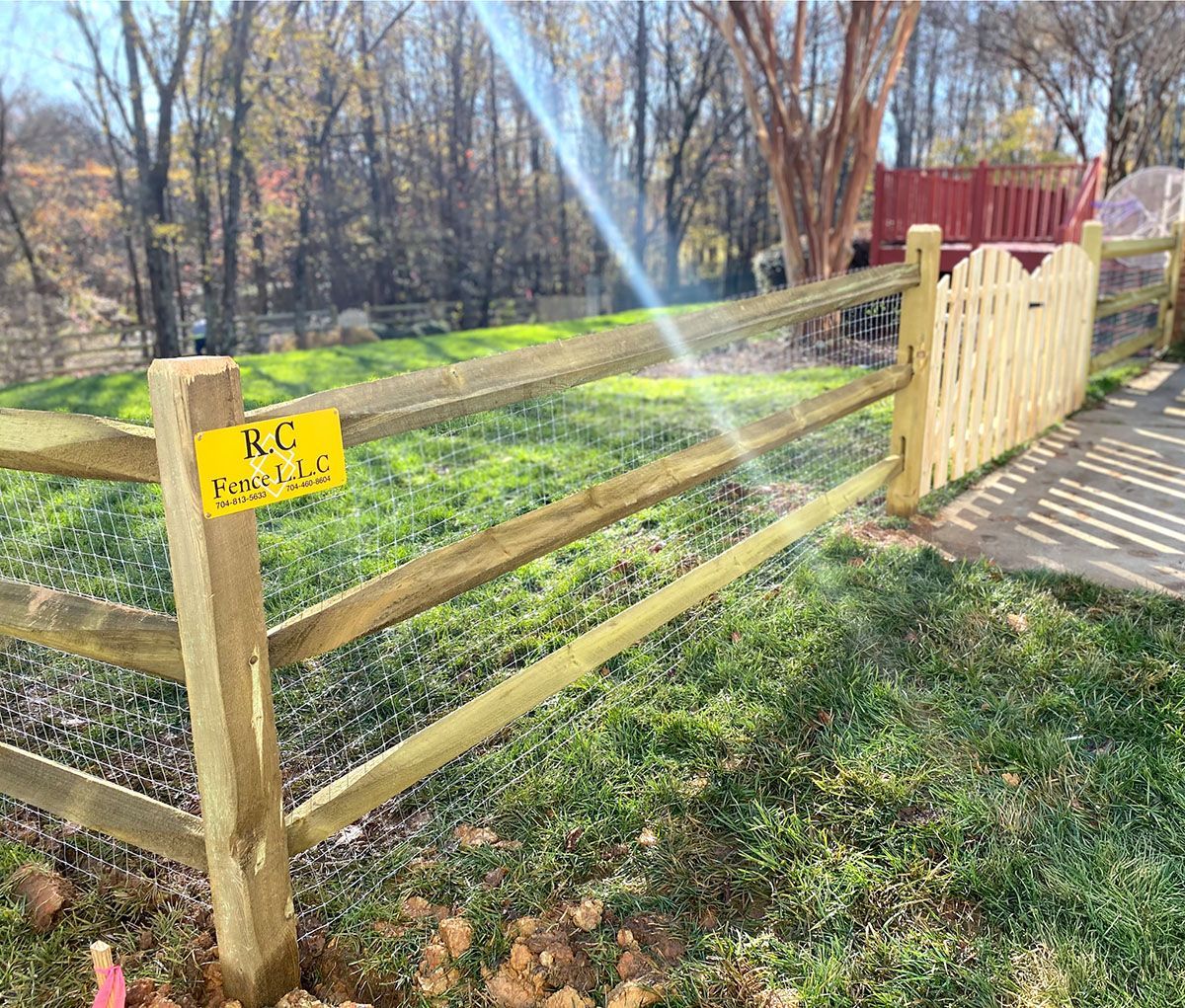 Wooden fence with wire mesh in a grassy yard, a yellow sign says “R.C. Regal Trails.” The sun shines through the trees.