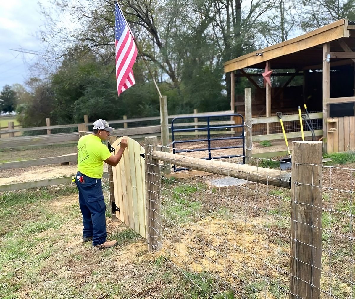 A man in a neon shirt installs a wooden fence gate in a rural area, with an American flag and a small building in the background.
