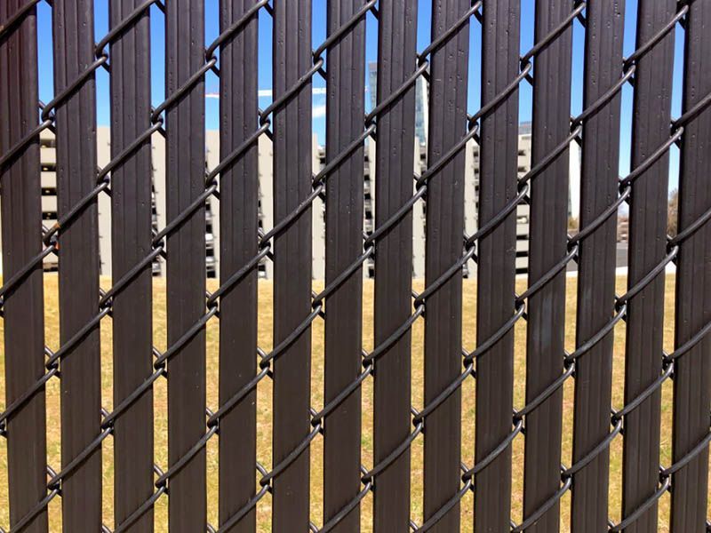 Brown privacy slats inserted into a chain link fence against a blue sky, partially obscuring a grassy area.