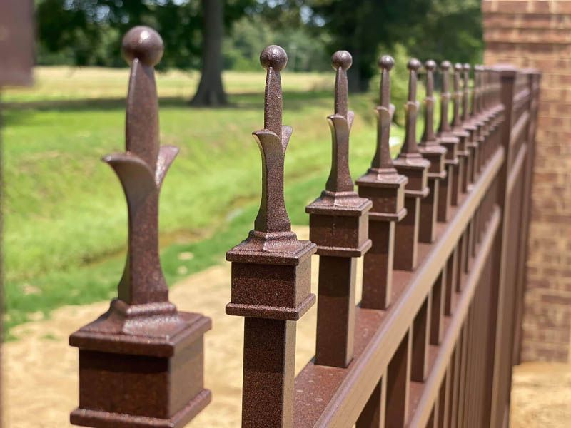 Brown metal fence with decorative finials, in front of a grassy area and brick wall.