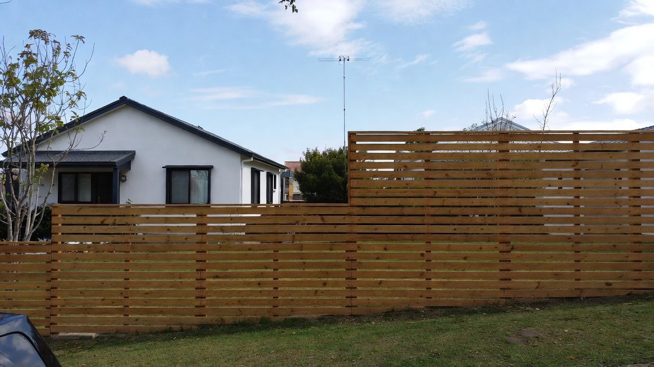A wooden slatted fence surrounds a white house with a dark roof against a cloudy sky.