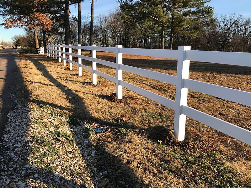 White three-rail fence along a roadside with shadows cast on brown grass.