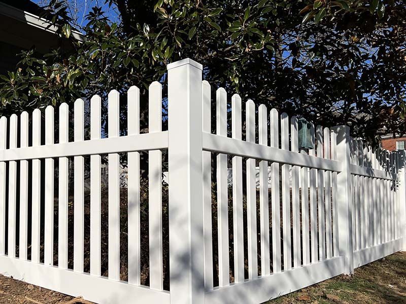 White picket fence in a yard, surrounding a corner. Fence has vertical pointed pickets and horizontal rails.