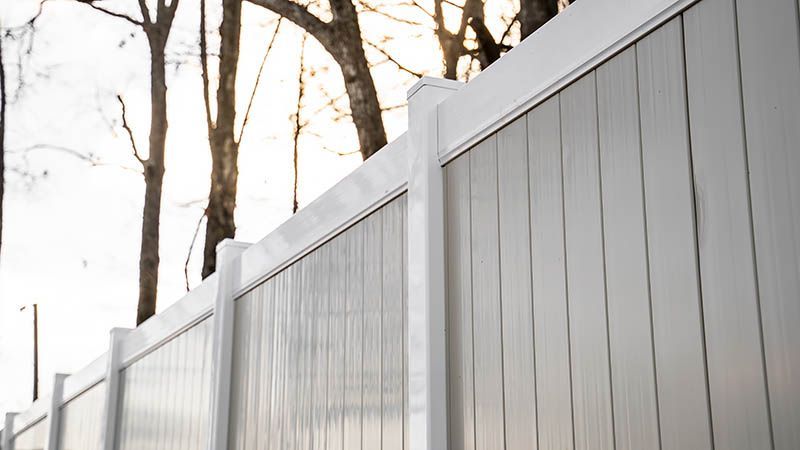 White vinyl privacy fence with vertical panels against a backdrop of trees and a bright sky.
