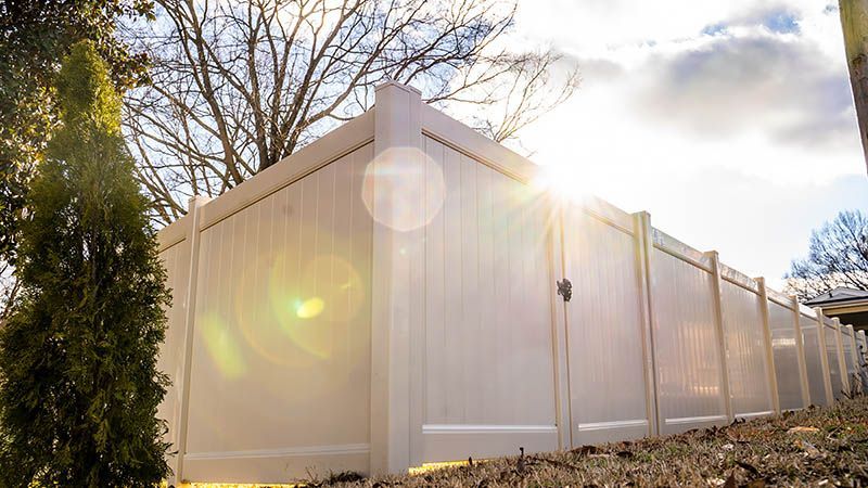 White vinyl fence with gate, sun shining through trees in the background.