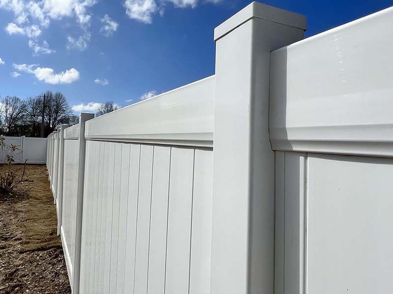 White vinyl fence against a blue sky with clouds. The fence is new and clean, and the setting appears to be a residential yard.
