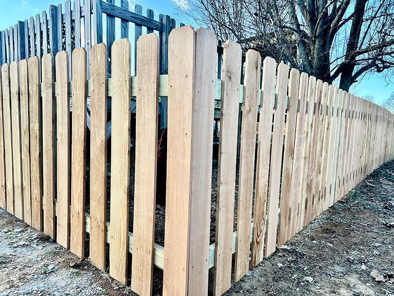 Wooden picket fence, constructed in a corner, extends towards the right. Pale brown wood, set outdoors.